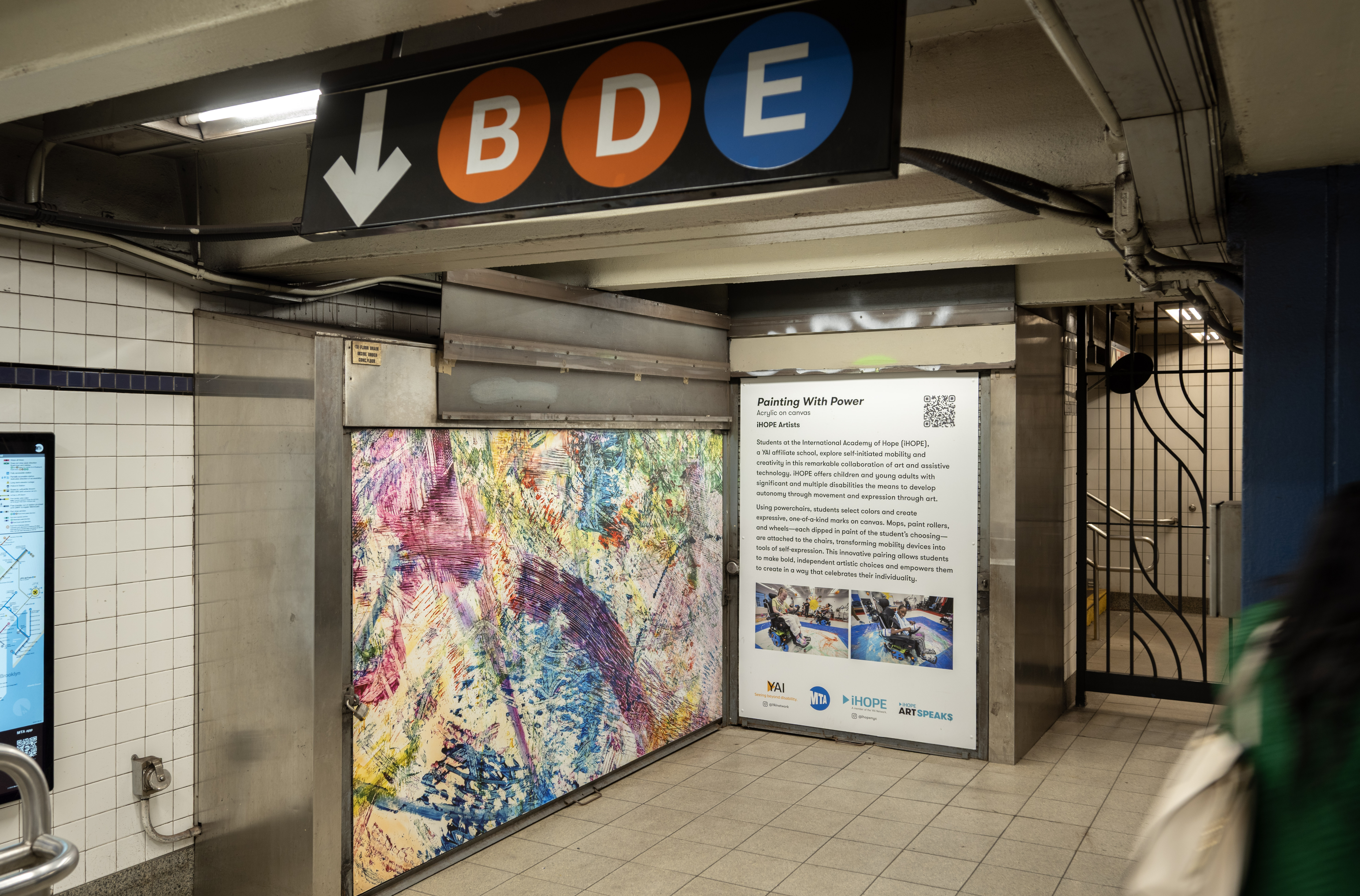 Abstract artwork printed on vinyl, displayed on a vacant unit in a subway station beneath signage for the B, D, and E trains. The piece is composed of layered, colorful paint textures in purple, pink, yellow, blue, and green, creating a dynamic composition.
