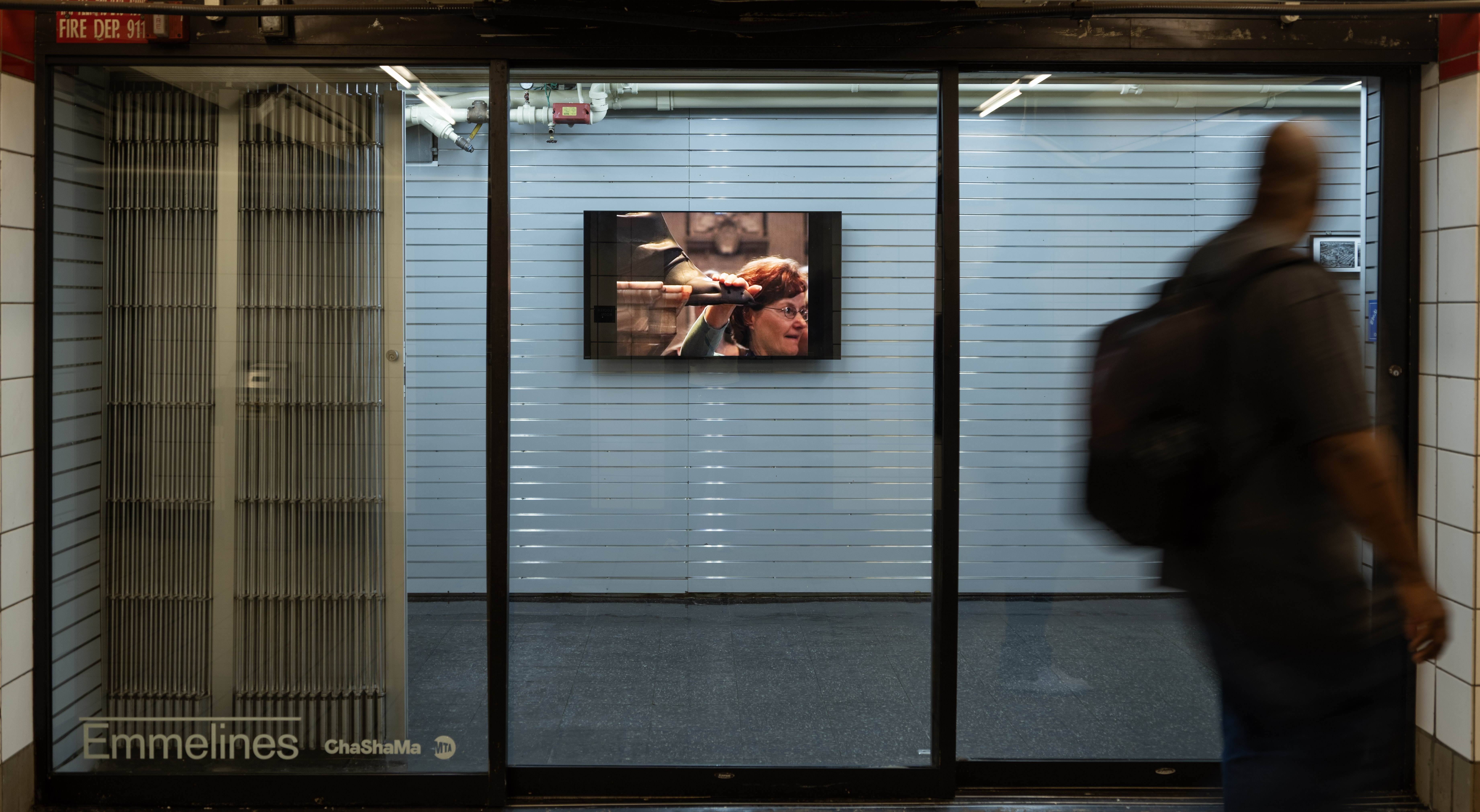Video artwork displayed on a TV inside a glass-fronted vacant retail unit. The still image on the screen shows a close-up of a woman with glasses partially obscured by a raised hand gripping a horizontal bar.